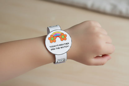 Child's hand wearing a colorful wristwatch with a rainbow and flower design on a green surface with pens.